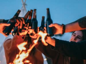 group of friends raising beer bottles by a camp fire